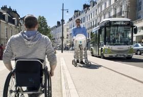 Une personne en fauteuil roulant croise une personne avec une poussette sur un trottoir accessible et doté d’une bande de guidage podotactile.