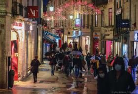 Nombreuses personnes masquées empruntant une rue commerçante sous les illuminations nocturnes de fin d’année.