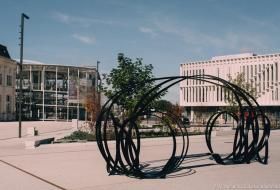 Sculpture de Pablo Reinoso sur le parvis de la gare. L’œuvre est faite de plusieurs tiges metalliques noires, qui forment deux ensembles de cercles reliés entre-eux, avec un banc au milieu. La rotonde et le bâtiment de l'Insa sont à l’arrière-plan. 