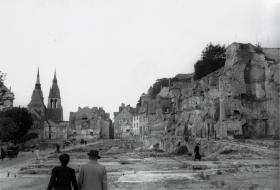 La place Louis 12 dévastée après les bombardements du 21 juin 1940. Les remparts du Château subsistent, ainsi que l’église Saint-Nicolas en arrière-plan. Deux personnes, de dos, s’avancent sur la place.