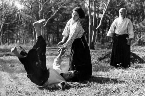 Une femme projette son adversaire au sol à l’aide d’une technique d’aïkido, sous l’œil d’un homme.