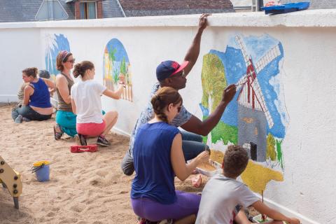 Des adultes et des enfants peignent une fresque sur le mur d’une aire de jeux extérieure.