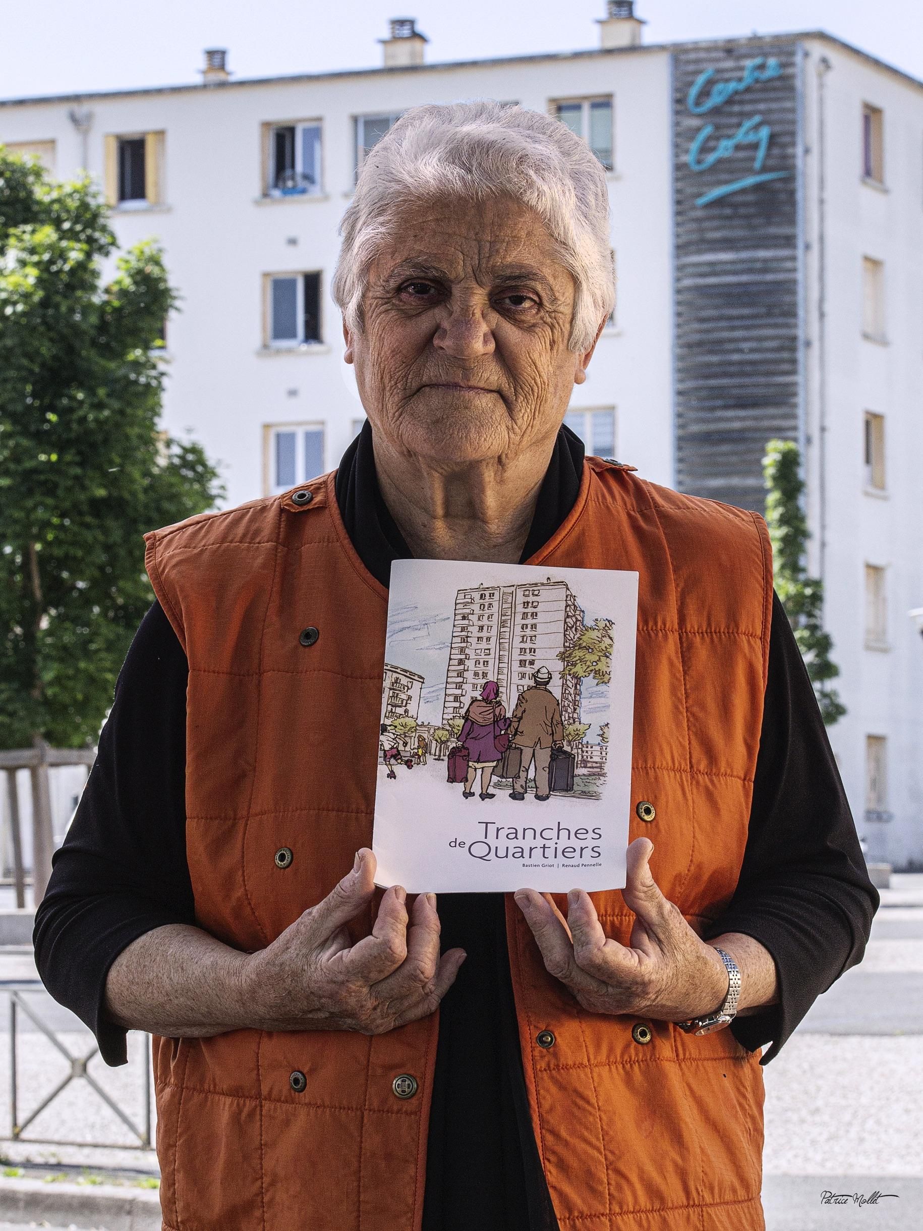 Adrienne Montesinos devant un immeuble du Centre Coty tient dans ses mains un livret « Tranches de quartier », dont la couverture face à l’objectif figure deux personnes avec leur valises face à un grand immeuble. 