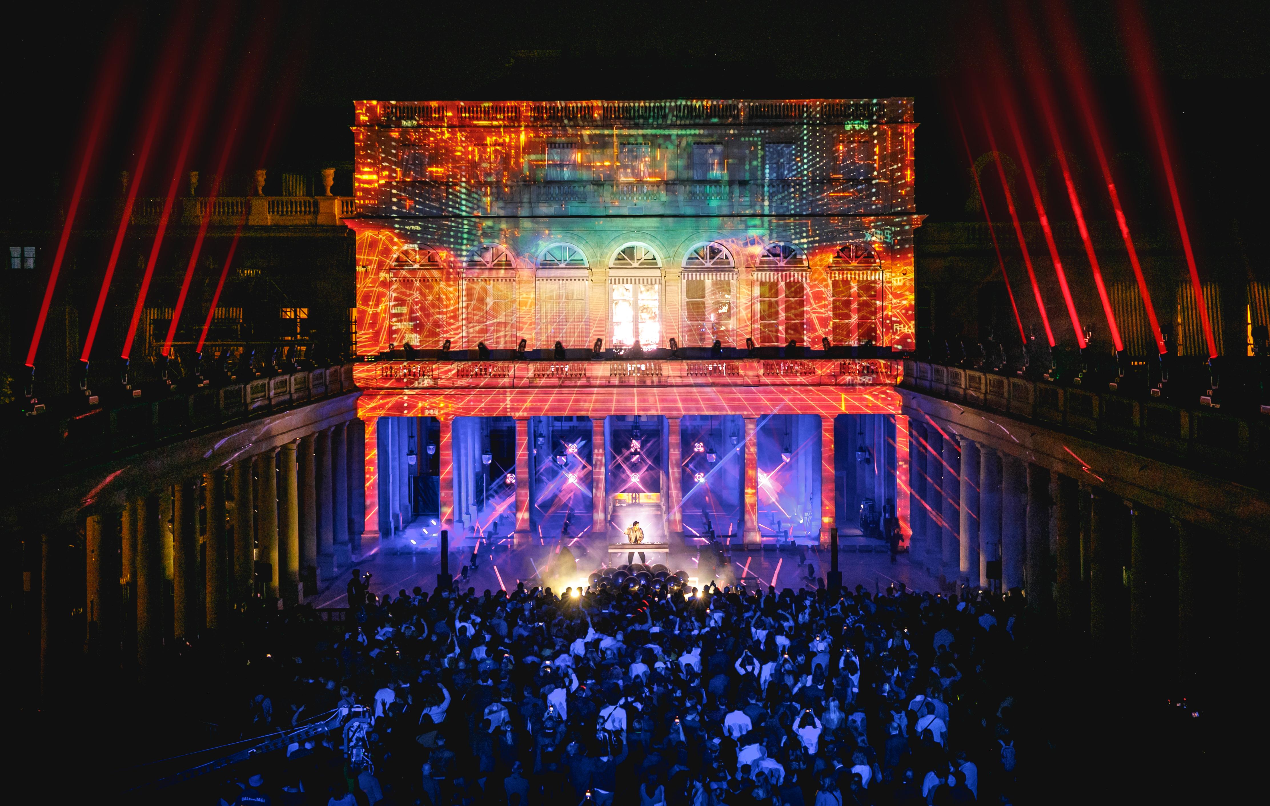 En plein spectacle, la foule dans la pénombre au premier plan, le DJ sous les projecteur, et les façades environnantes éclairées par de nombreux lasers de toutes les couleurs.