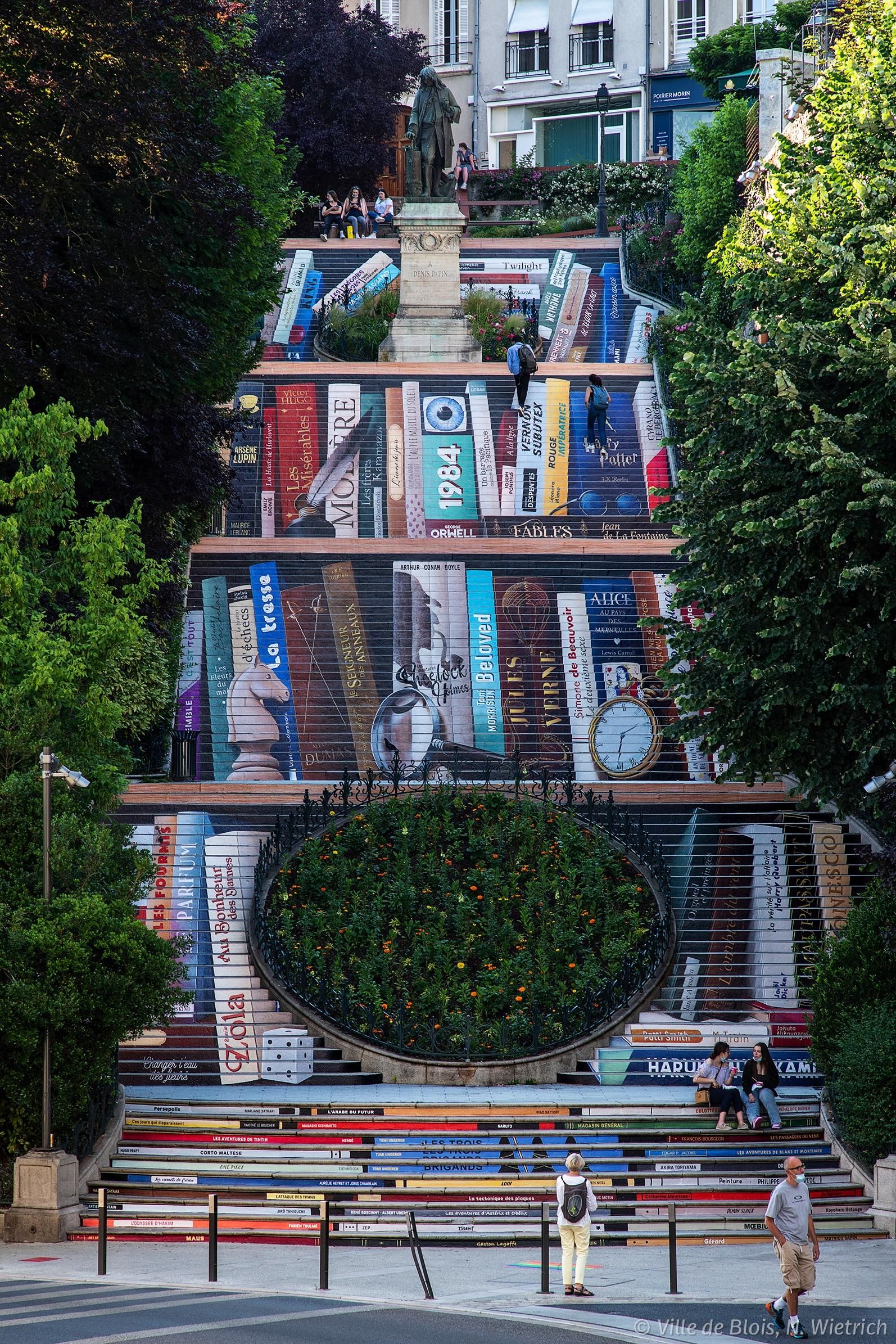 Photo d’ensemble de l’escalier, dont les contremarches sont décorées pour ensemble former l’illusion d’une bibliothèque géante, comportant une centaine de livres.