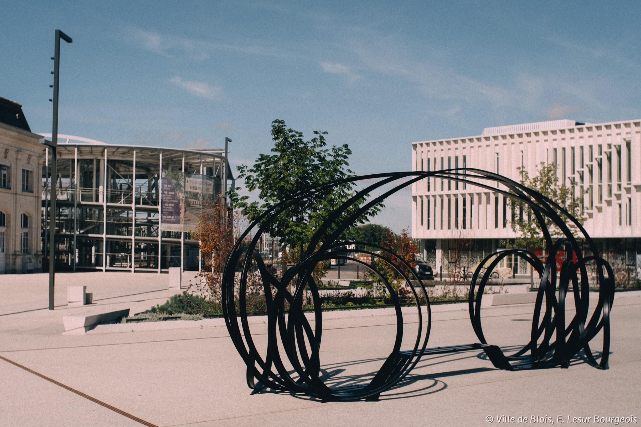 Sculpture de Pablo Reinoso sur le parvis de la gare. L’œuvre est faite de plusieurs tiges metalliques noires, qui forment deux ensembles de cercles reliés entre-eux, avec un banc au milieu. La rotonde et le bâtiment de l'Insa sont à l’arrière-plan. 